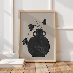 May include: A framed art print featuring a black vase with two handles and a textured surface. The vase holds black floral stems against a beige background. The frame is light wood, and the artwork is displayed on a wooden floor.