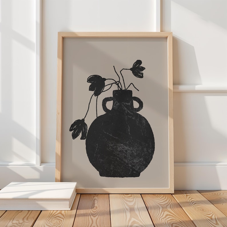 May include: A framed art print featuring a black vase with two handles and a textured surface. The vase holds black floral stems against a beige background. The frame is light wood, and the artwork is displayed on a wooden floor.