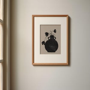 May include: A framed art print featuring a black vase with flowers. The vase has a textured appearance and is set against a neutral background. The artwork is enclosed in a light brown wooden frame, suitable for home decor.
