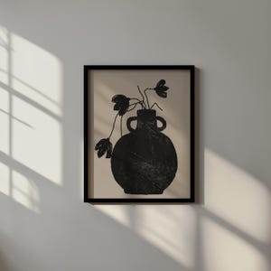 May include: Black framed art print featuring a black vase with two handles, holding black flowers against a beige background. The vase has a textured appearance. The artwork is displayed on a white wall with shadows.