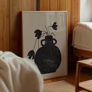 May include: A framed art print featuring a black vase with flowers against a beige background. The vase has two handles and the flowers have a simple, abstract design. The frame is a light wood color, and the artwork is displayed in a room with wooden paneling.