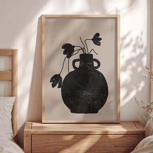 May include: A framed art print featuring a black vase with two handles, holding black floral stems against a beige background. The artwork is in a wooden frame, creating a minimalist aesthetic. The print is displayed on a wooden nightstand.