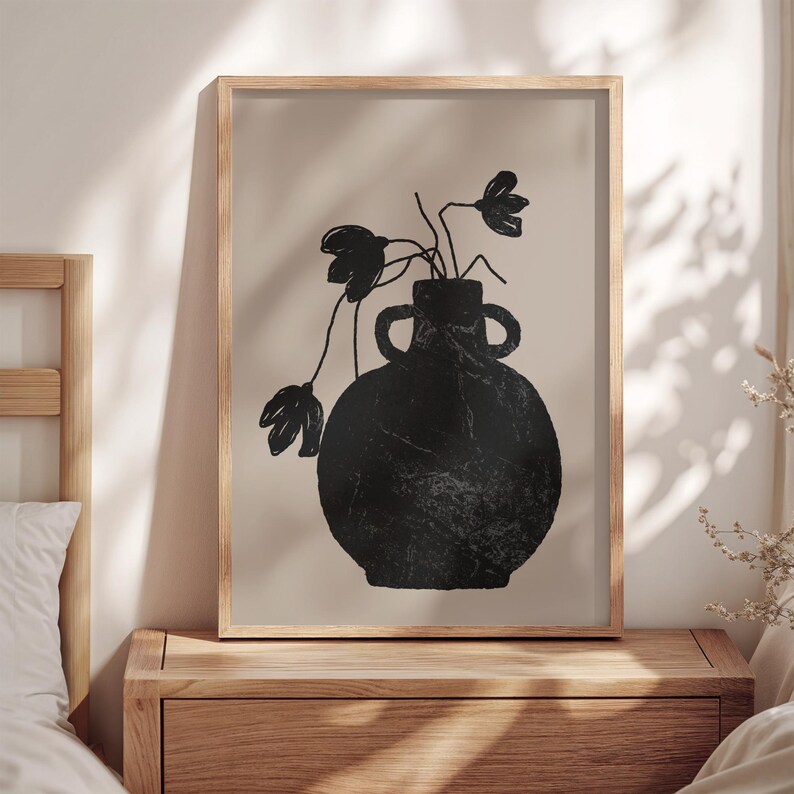 May include: A framed art print featuring a black vase with two handles, holding black floral stems against a beige background. The artwork is in a wooden frame, creating a minimalist aesthetic. The print is displayed on a wooden nightstand.