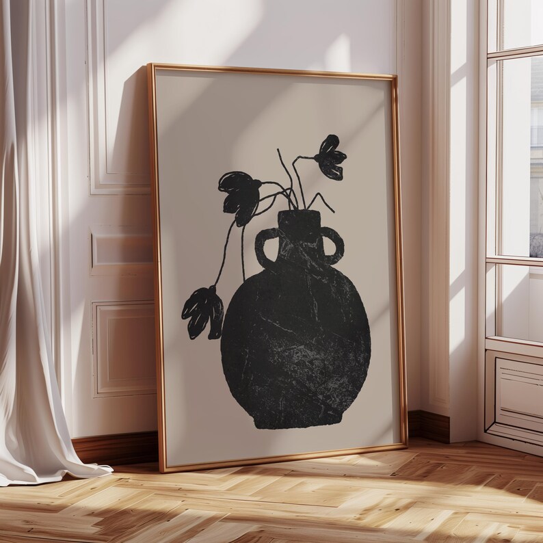 May include: A framed art print featuring a black vase with flowers against a beige background. The vase has two handles and a textured appearance. The artwork is in a gold frame, leaning against a white wall and wooden floor.