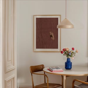 K&ouml;nnte beinhalten: Ein gerahmter Druck mit braunem Hintergrund und abstrakten Linien h&auml;ngt an einer wei&szlig;en Wand. Eine blaue Vase mit Blumen steht auf einem runden Tisch mit einem Holzstuhl. Eine beige Pendelleuchte h&auml;ngt &uuml;ber dem Tisch.