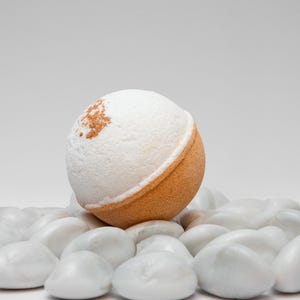 May include: A spherical bath bomb with a white, textured top and a tan base, lightly dusted with brown powder. It sits on a bed of smooth, white stones, evoking a spa atmosphere.