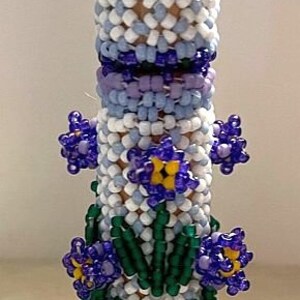 Peut inclure: Un bouchon de bouteille en perles avec une base blanche et bleue, des feuilles vertes et des fleurs violettes avec des centres jaunes.