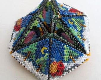 Puede incluir: Un adorno de cuentas geométrico multifacético con un diseño en forma de estrella. El adorno está hecho de cuentas de colores en tonos de azul, verde, rojo y blanco. Las cuentas están dispuestas en un patrón complejo que crea un efecto tridimensional.
