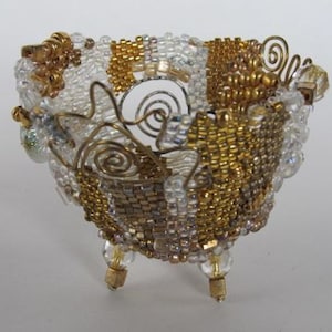 May include: A small, gold and white beaded bowl with a wire frame and three legs. The bowl is made of small, iridescent beads and has a delicate, intricate design.