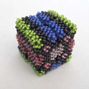 Può includere: Un piccolo cubo colorato fatto di perline. Il cubo è fatto di perline nere, verdi, blu, rosa e bianche. Le perline sono disposte in uno schema geometrico.