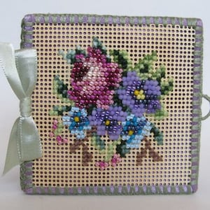 May include: A small, square, purple and green needlepoint canvas with a floral design. The design features a bouquet of purple, pink, and blue flowers with green leaves. The canvas is framed with a purple and green border.