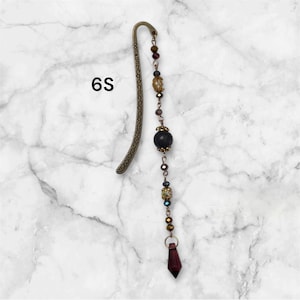 May include: A bronze bookmark with a decorative design and a chain of beads with a black bead and a burgundy crystal at the end. The bookmark is labeled "6S".