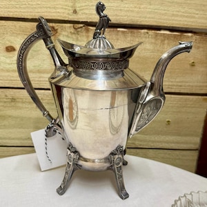 May include: A silver-colored teapot with a decorative handle and spout. The teapot has a lion figure on the lid and rests on three ornate legs. The teapot is displayed on a white surface against a wooden background.