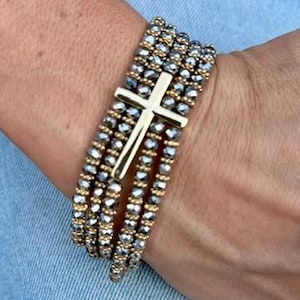 May include: A beaded bracelet with a gold cross charm. The bracelet features multiple strands of gray and gold beads. The cross is centered on the bracelet and has a polished finish. The bracelet is shown on a wrist against a denim background.