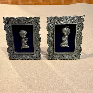 May include: Two ornate, silver-colored picture frames. Each frame has a dark blue background and features a small, silver-colored sculpture of a child kneeling in prayer. The frames have detailed, scrolling designs.