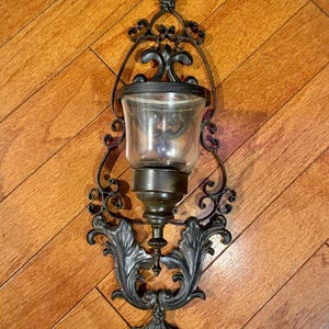 Puede incluir: Aplique de pared de metal negro ornamentado con un portavelas de cristal transparente. El aplique presenta volutas decorativas y un diseño de flor de lis en la parte superior. Montado sobre una superficie de madera.