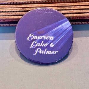 Op de afbeelding: Een ronde, paarse button met de tekst "Emerson Lake & Palmer" in witte letters. De button heeft een blauw en wit straalontwerp en de tekst "...only to the show..." bovenaan. De button ligt op een grijze ondergrond.