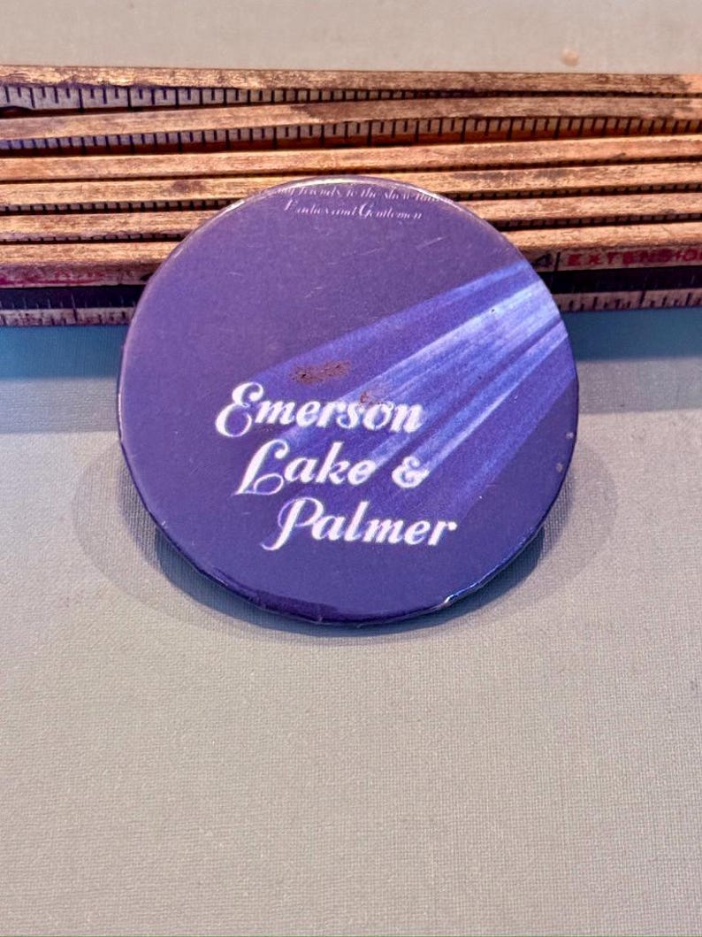 Op de afbeelding: Een ronde, paarse button met de tekst "Emerson Lake & Palmer" in witte letters. De button heeft een blauw en wit straalontwerp en de tekst "...only to the show..." bovenaan. De button ligt op een grijze ondergrond.