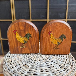 May include: Two wooden bookends with a rooster illustration. The bookends are arched at the top and feature a yellow rooster with red comb and green tail feathers. The bookends are made of a medium-toned wood and are displayed on a woven basket.