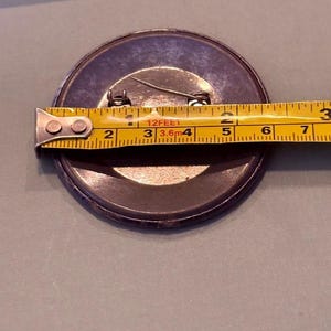 Op de afbeelding: Een grote, ronde knoop met een speld, met een diameter van ongeveer 7,6 cm. De knoop is donker met een metalen rand en een gele meetlint ligt eroverheen.