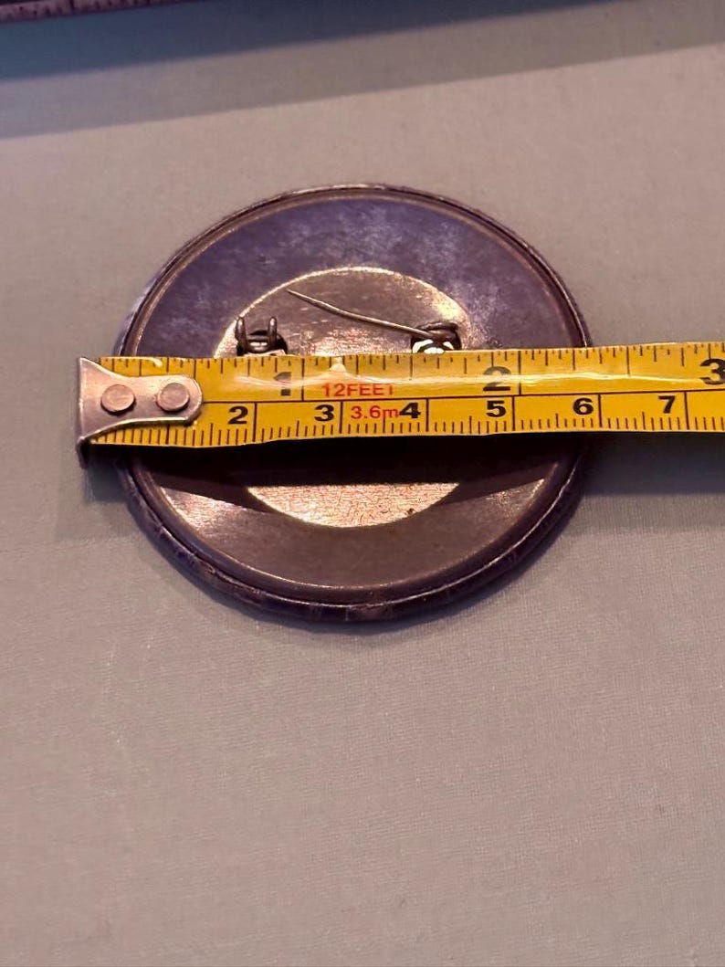 Op de afbeelding: Een grote, ronde knoop met een speld, met een diameter van ongeveer 7,6 cm. De knoop is donker met een metalen rand en een gele meetlint ligt eroverheen.