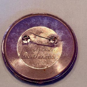 Op de afbeelding: Een ronde, paars getinte knoopspeld met een veiligheidsspeld. De speld heeft een metallic afwerking en bevat de tekst "BADGE LASALLE" en "MINIT ILL 61301". De speld is waarschijnlijk een promotieartikel of verzamelobject.