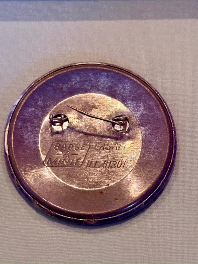 Op de afbeelding: Een ronde, paars getinte knoopspeld met een veiligheidsspeld. De speld heeft een metallic afwerking en bevat de tekst "BADGE LASALLE" en "MINIT ILL 61301". De speld is waarschijnlijk een promotieartikel of verzamelobject.