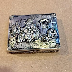 May include: A rectangular silver-toned box with a detailed relief scene of people and buildings. The relief depicts figures in period clothing, a house, and a dog. The box has ornate edges and a hinged lid, suggesting it may be a trinket box or similar item.