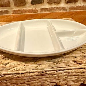 Könnte beinhalten: Eine weiße, ovale Servierschale mit drei Fächern. Die Schale steht auf einem gewebten, rechteckigen Platzset. Der Hintergrund zeigt eine Ziegelmauer und eine Holzoberfläche.