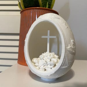 Pode incluir: Um objeto decorativo branco em forma de ovo com uma frente aberta, apresentando uma cruz no interior e pequenas pedras brancas na base. O ovo tem um design em relevo detalhado. Um vaso laranja com uma planta verde e amarela está no fundo.
