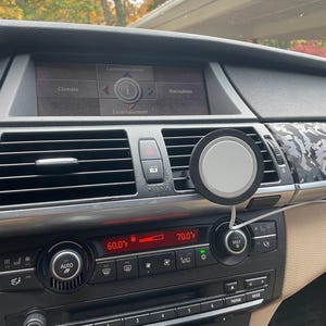 Op de afbeelding: Auto-dashboard met een digitaal display met klimaat- en navigatie-opties. Een ronde, grijze telefoonlader is aan het dashboard bevestigd. De temperatuurweergave toont 15.6°C en 21.1°C.