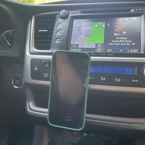 Può includere: Uno smartphone con bordo verde è montato sul cruscotto di un'auto. Lo schermo di infotainment dell'auto mostra una mappa e informazioni musicali. Il cruscotto ha vari pulsanti e bocchette. La temperatura dell'auto è impostata a 20 gradi Celsius.