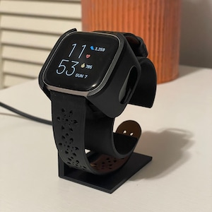 May include: A black smartwatch with a square face displaying the time as 11:53, with a black band and a decorative cutout design. The watch is on a black stand with a brown leather accent. The watch shows 3,259 steps and 785 calories burned.