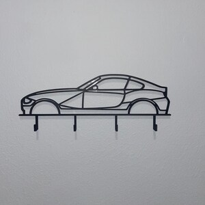 May include: Black metal key holder shaped like a sports car. The key holder has four hooks below the car silhouette. The car design includes the wheels, windows, and body lines. The key holder is mounted on a white wall.