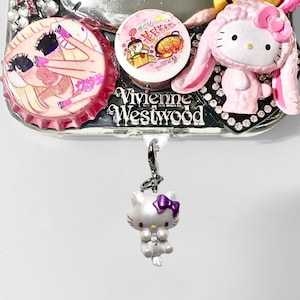 May include: A silver-colored Hello Kitty charm with a purple bow hangs below a decorative piece featuring a pink bottle cap with a cartoon image, a round sticker, and a pink Hello Kitty figure with bunny ears. The word "Vivienne Westwood" is visible.