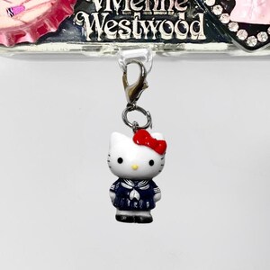 May include: A silver phone case adorned with multiple decorative items. It includes a Hello Kitty charm, a pink and white bottle cap design, and a Vivienne Westwood logo. The Hello Kitty charm is dressed in a sailor suit.