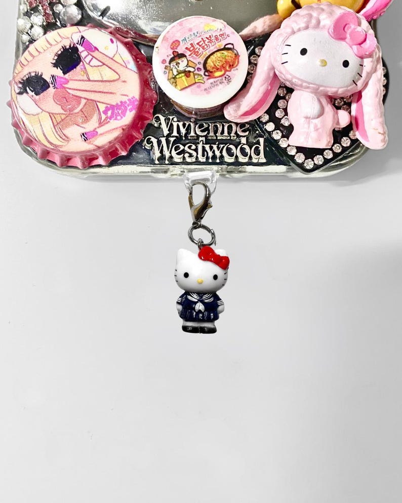 May include: A silver phone case adorned with multiple decorative items. It includes a Hello Kitty charm, a pink and white bottle cap design, and a Vivienne Westwood logo. The Hello Kitty charm is dressed in a sailor suit.