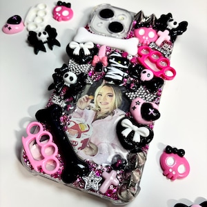 Puede incluir: Una funda de teléfono transparente decorada con dijes de resina rosas y negros que incluyen calaveras, lazos, huesos y puños americanos. El centro presenta una foto de una persona con un recipiente de comida, cubierta de brillantes rosas y plateados.