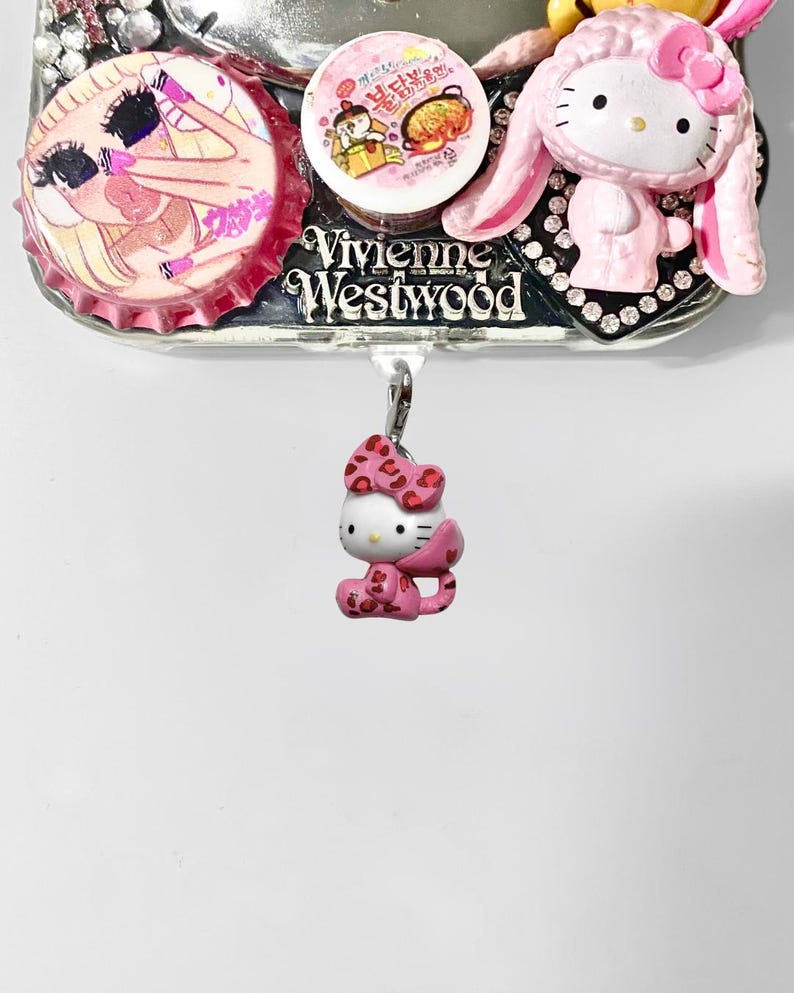 May include: A decorative phone case with various charms. It features a pink Hello Kitty figure, a bottle cap with a cartoon illustration, and a Vivienne Westwood logo. The charms are attached to a silver base.