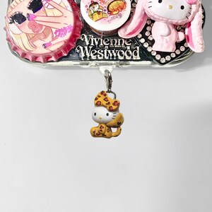 May include: A decorative phone case with a variety of charms. The case features a Hello Kitty charm in a leopard print outfit, a pink bunny-eared Hello Kitty, and a Vivienne Westwood logo. Additional charms include bottle caps with illustrations.