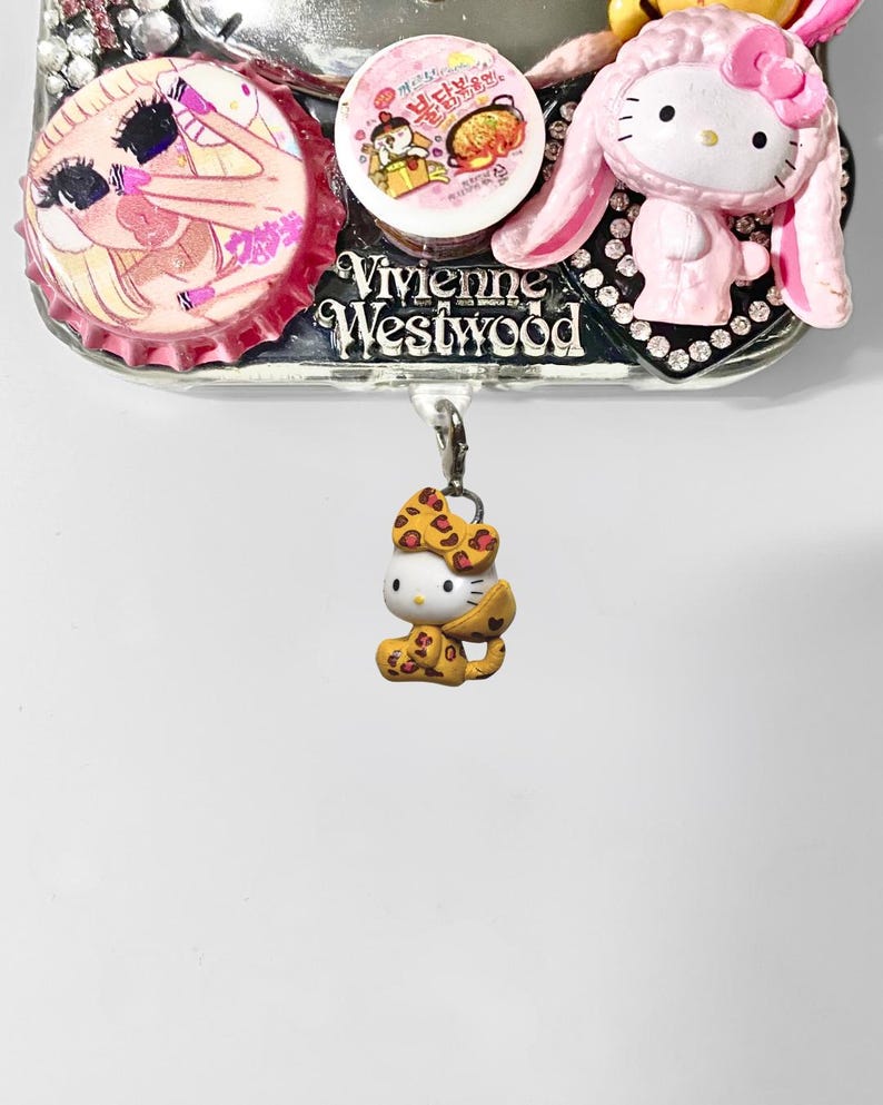 May include: A decorative phone case with a variety of charms. The case features a Hello Kitty charm in a leopard print outfit, a pink bunny-eared Hello Kitty, and a Vivienne Westwood logo. Additional charms include bottle caps with illustrations.