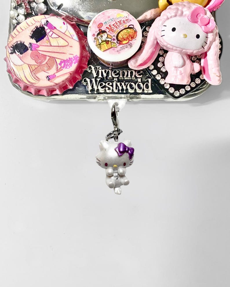 May include: A silver-colored Hello Kitty charm with a purple bow hangs below a decorative piece featuring a pink bottle cap with a cartoon image, a round sticker, and a pink Hello Kitty figure with bunny ears. The word "Vivienne Westwood" is visible.