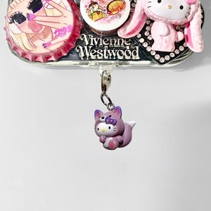 May include: A silver-toned Vivienne Westwood charm with a purple Hello Kitty fox figurine hanging from a chain. The charm also features a pink Hello Kitty in a bunny costume, and two bottle cap designs.