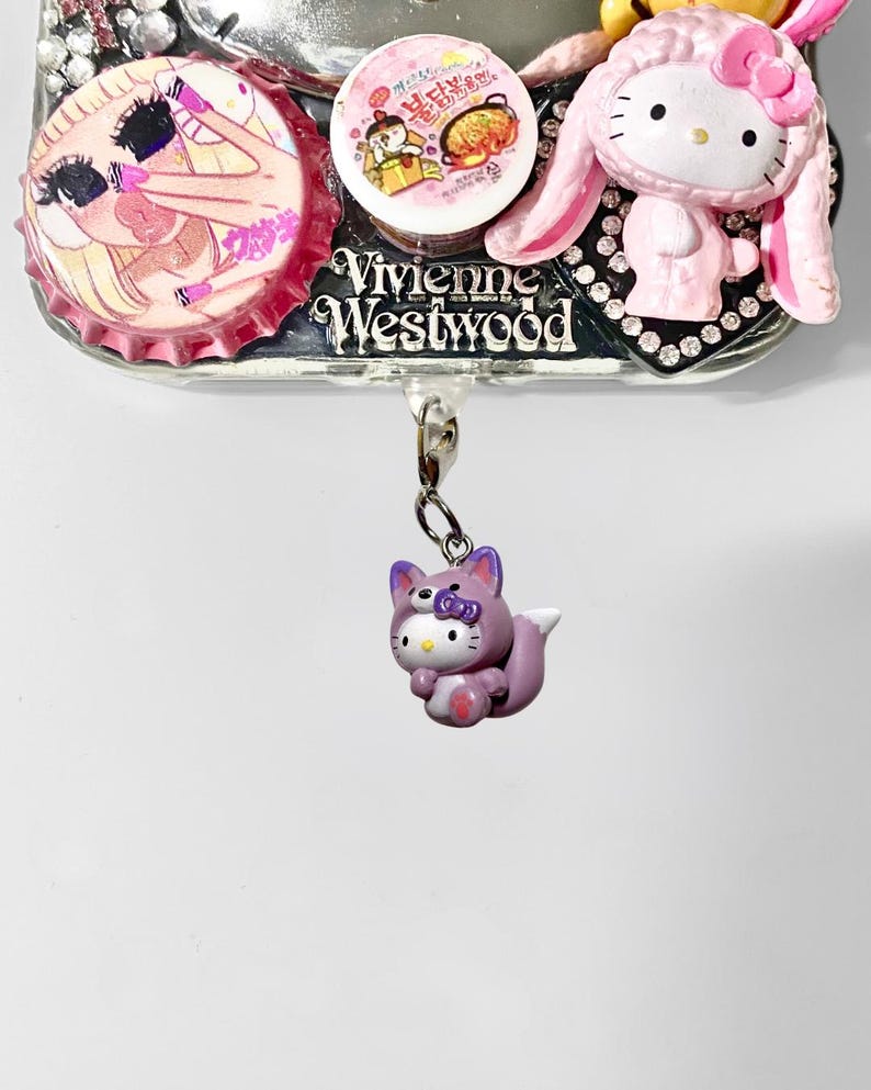 May include: A silver-toned Vivienne Westwood charm with a purple Hello Kitty fox figurine hanging from a chain. The charm also features a pink Hello Kitty in a bunny costume, and two bottle cap designs.