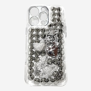 Puede incluir: Funda de teléfono transparente con adornos plateados y grises. La funda presenta un candado en forma de corazón, un corazón cubierto de pedrería y otros abalorios decorativos. La funda está cubierta de tachuelas redondas grises.