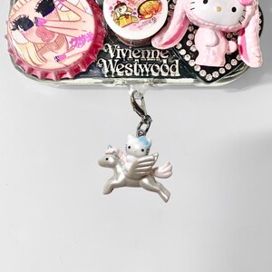 May include: A decorative charm featuring a Hello Kitty figure riding a pegasus. The charm is attached to a silver-colored metal base with other decorative elements, including a pink bottle cap and a Hello Kitty figure in a bunny costume.