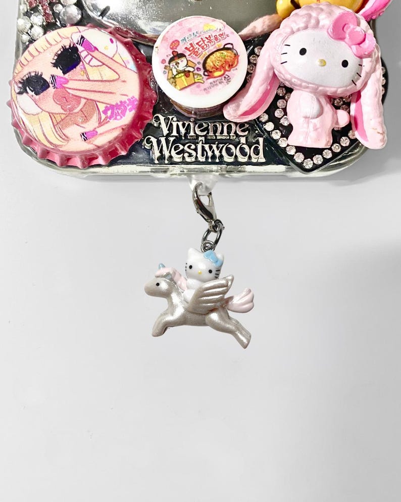 May include: A decorative charm featuring a Hello Kitty figure riding a pegasus. The charm is attached to a silver-colored metal base with other decorative elements, including a pink bottle cap and a Hello Kitty figure in a bunny costume.