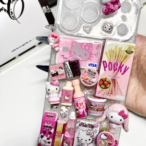 May include: A clear phone case decorated with miniature 3D charms including a Hello Kitty figure, a strawberry Pocky box, a pink Visa card, a bottle of sauce, a face pack, and various food-themed items. The charms are arranged across the back surface.