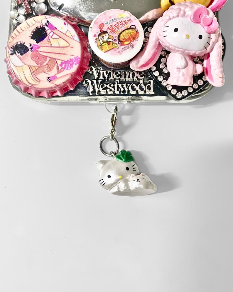 May include: A decorative phone case with various charms. The case features a pink Hello Kitty figure with bunny ears, a bottle cap with a cartoon illustration, and a small white cat charm. The case has the text "Vivienne Westwood".