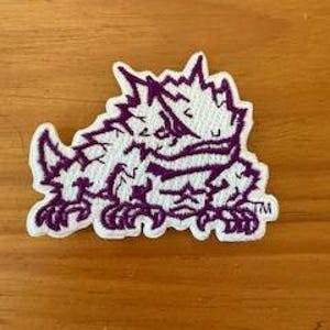 May include: Embroidered patch featuring a purple and white horned lizard design. The patch has a detailed outline and is sewn onto a white background. The lizard is in a crouched position, with the trademark symbol visible.
